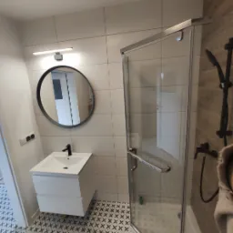 Nowoczesna łazienka z kabiną prysznicową ze srebrnymi okuciami, okrągłym lustrem z czarną ramą nad białą szafką z umywalką i wzorzystymi, czarno-białymi płytkami podłogowymi.