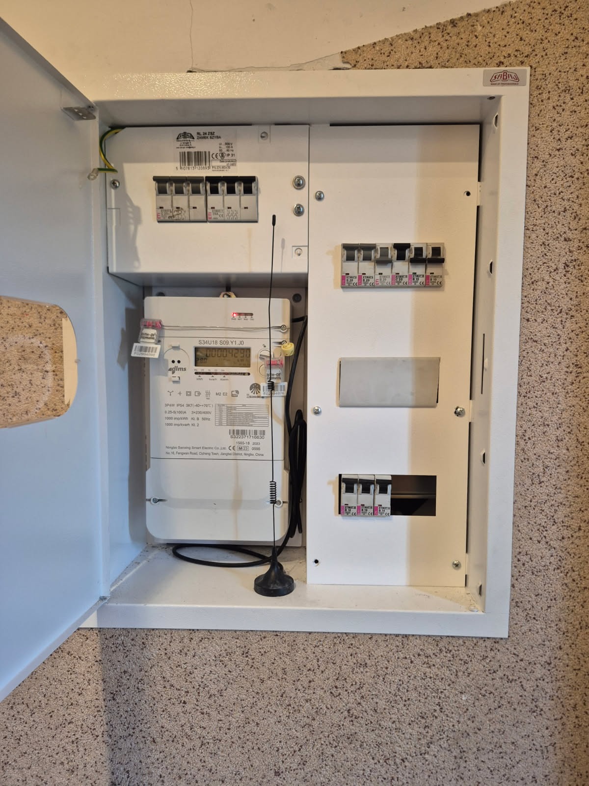 Otwarta skrzynka elektryczna z licznikiem i bezpiecznikami, widok z bliska. Biała obudowa na beżowej ścianie. Widoczny numer licznika: 00004290.