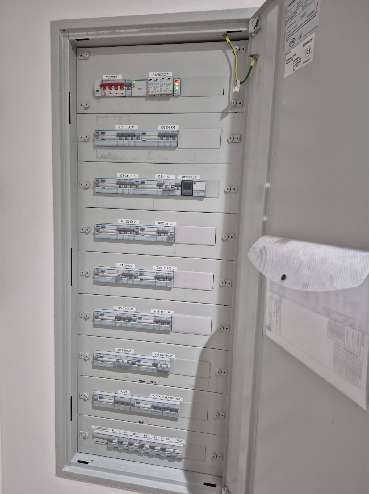 Otwarta skrzynka elektryczna z bezpiecznikami i oznaczeniami, widok z bliska. Widoczne przewody i dokumentacja wewnątrz. Szara obudowa.