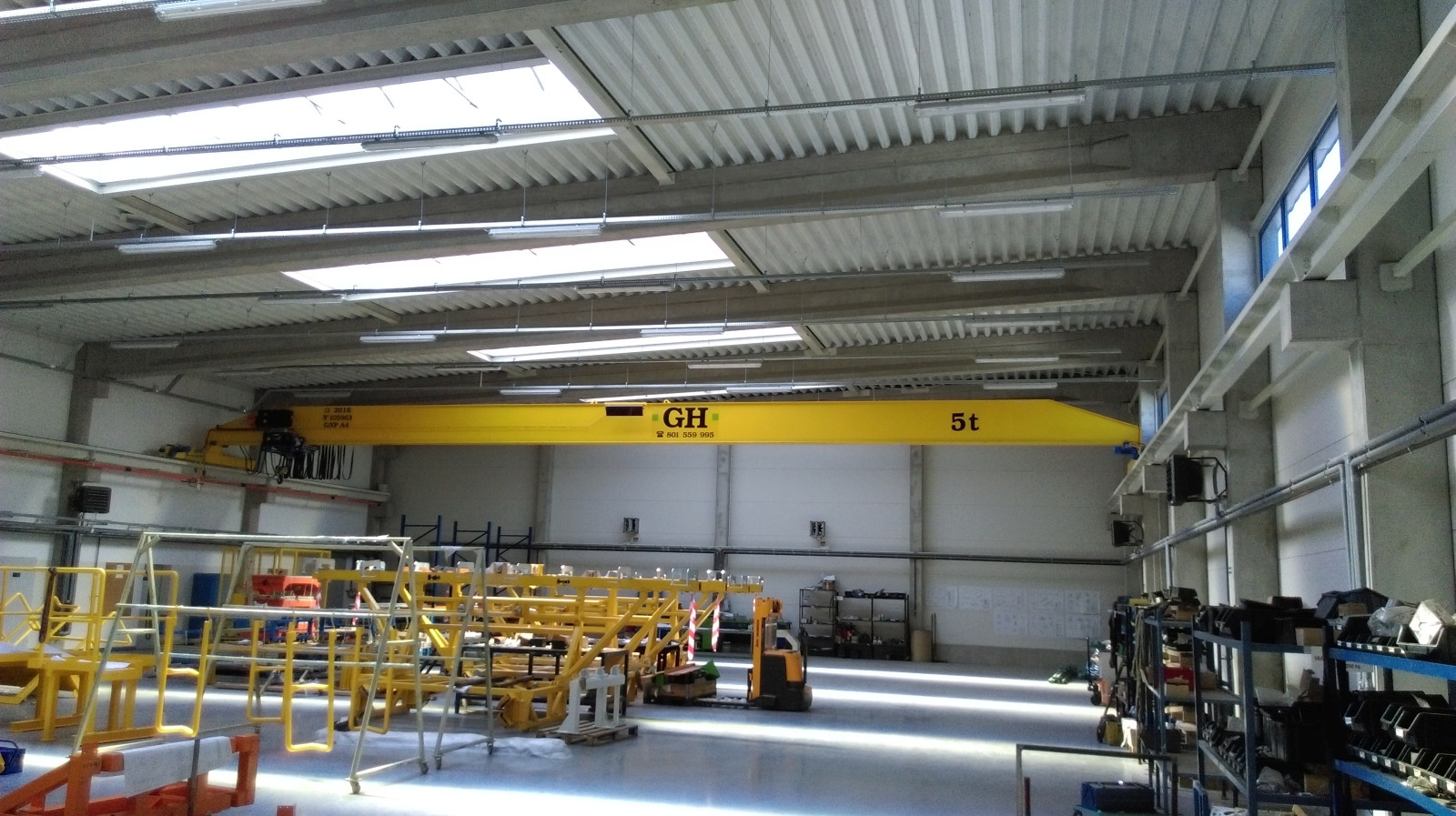 Wnętrze hali produkcyjnej z żółtym suwnicą bramową o udźwigu 5 ton, oświetlonej świetlikami dachowymi i lampami, z widocznym osprzętem i elementami konstrukcji stalowej.
