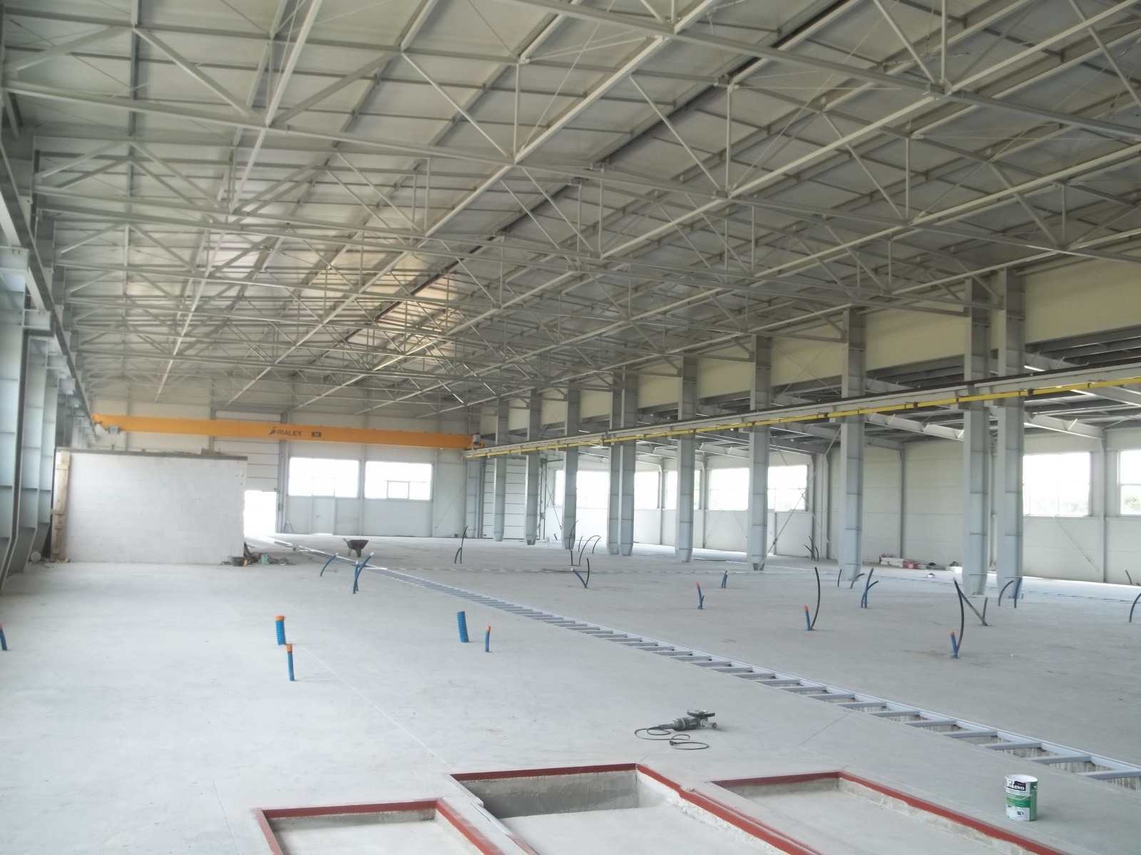 Wnętrze hali w stanie surowym, widoczne stalowe elementy konstrukcyjne dachu, słupy, okna, instalacje w podłodze oraz suwnica pod stropem.