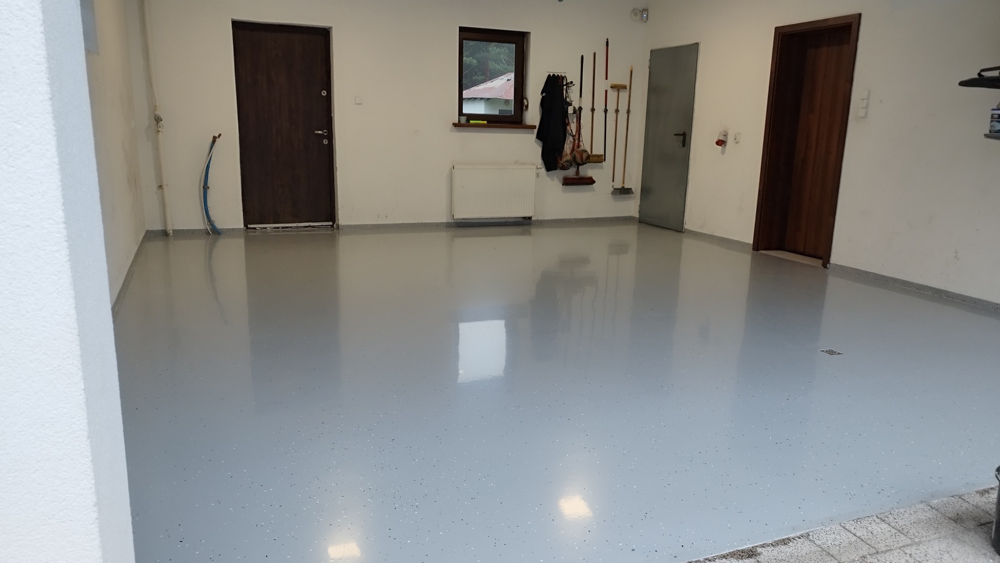 Jasnoszara posadzka z drobnymi, ciemniejszymi plamkami w garażu, odbijająca światło, z widocznymi drzwiami, oknem i narzędziami na ścianie.