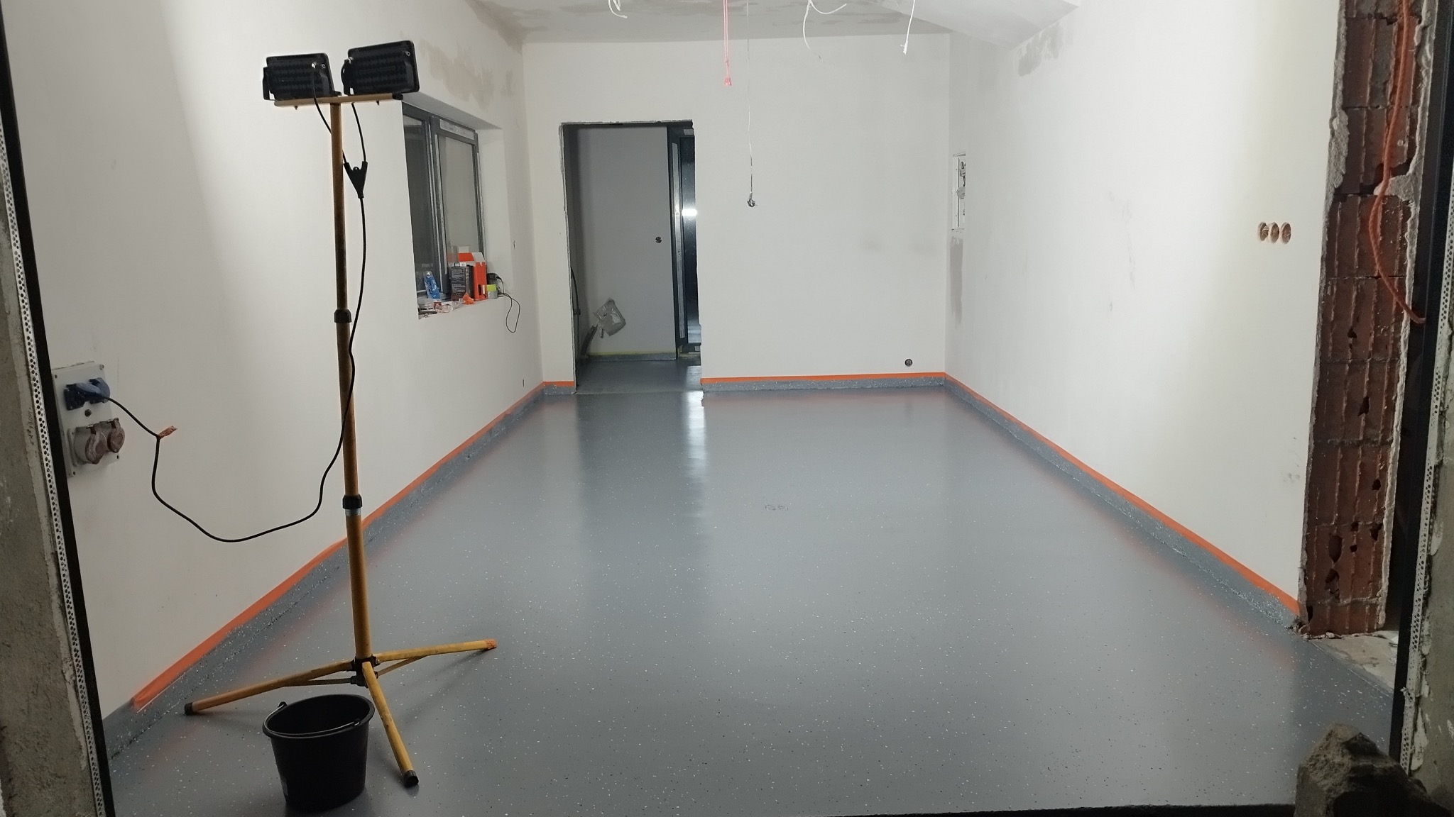 Szara, błyszcząca posadzka z drobnymi kolorowymi elementami, zabezpieczona pomarańczową taśmą malarską przy ścianach, oświetlona lampą budowlaną z wiadrem u podstawy.