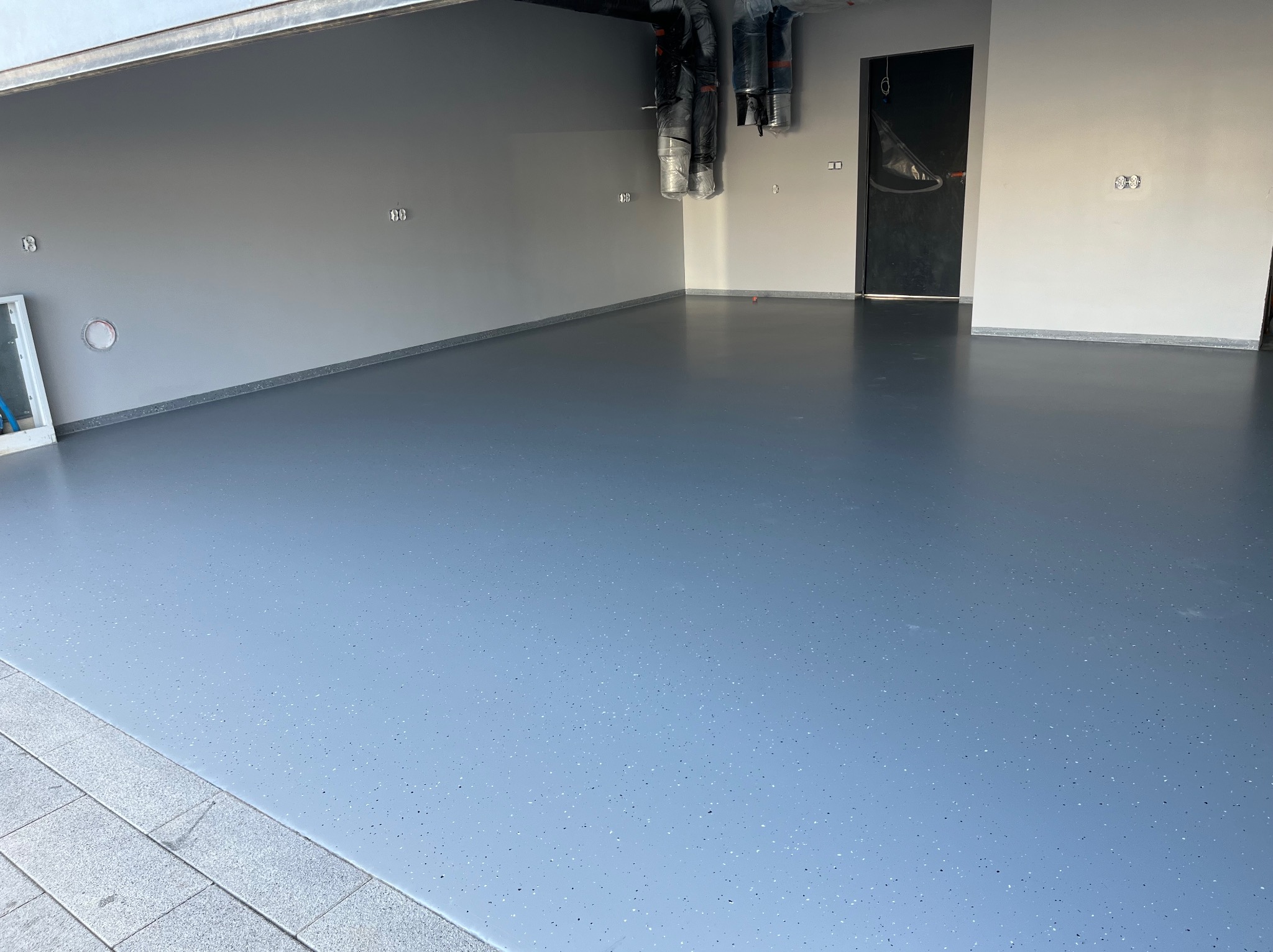 Jasnoszara, gładka podłoga z delikatnym wzorem drobnych kropek, świeżo wylana w pustym garażu z szarymi ścianami i czarnymi drzwiami, widoczne zabezpieczenia na ścianach.
