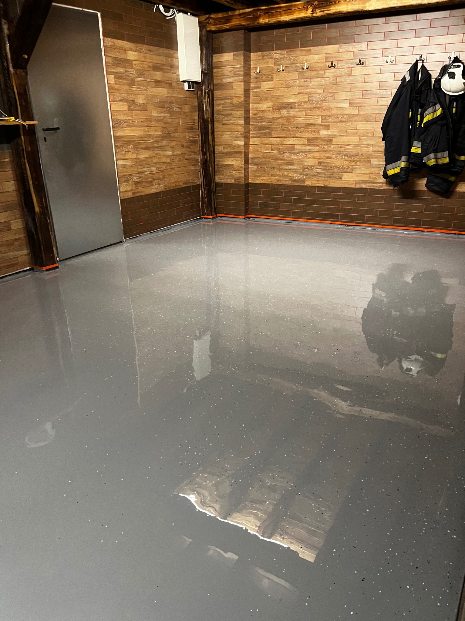 Błyszcząca, szara posadzka z drobnymi, ciemnymi inkluzjami w pomieszczeniu z brązowymi, drewnopodobnymi płytkami na ścianach i wiszącymi ubraniami roboczymi strażaka.
