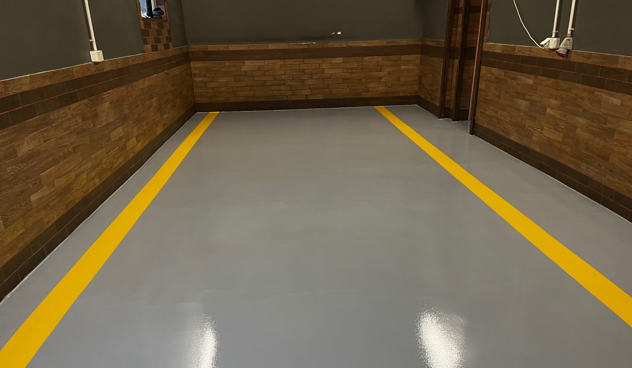 Szara, błyszcząca posadzka z wyraźnymi żółtymi liniami wyznaczającymi strefy, otoczona ścianami z dekoracyjną okładziną imitującą cegłę.