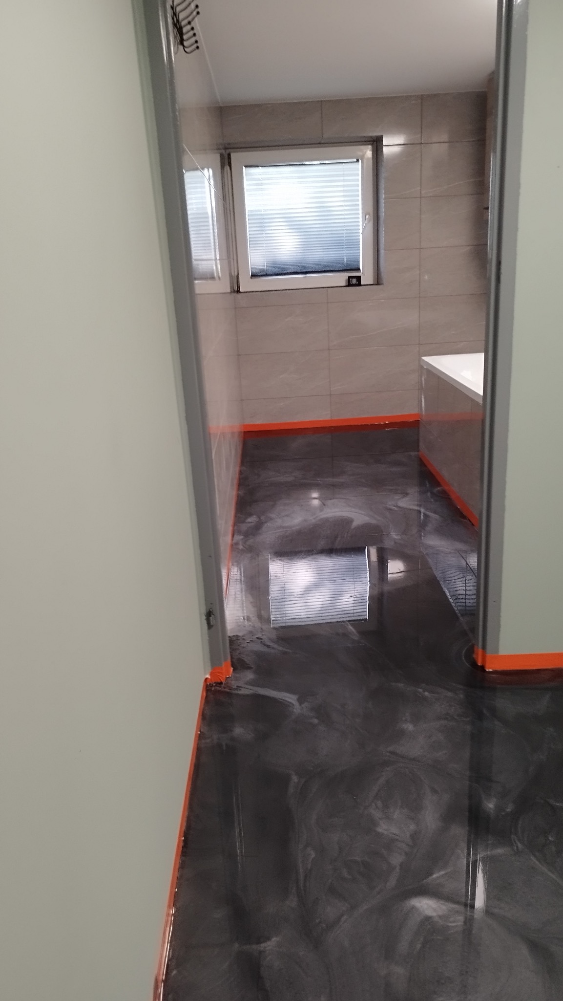 Błyszcząca, ciemnoszara posadzka żywiczna z marmurkowym wzorem w łazience, zabezpieczona pomarańczową taśmą malarską przy ścianach i framugach drzwi, z widocznym odbiciem okna.
