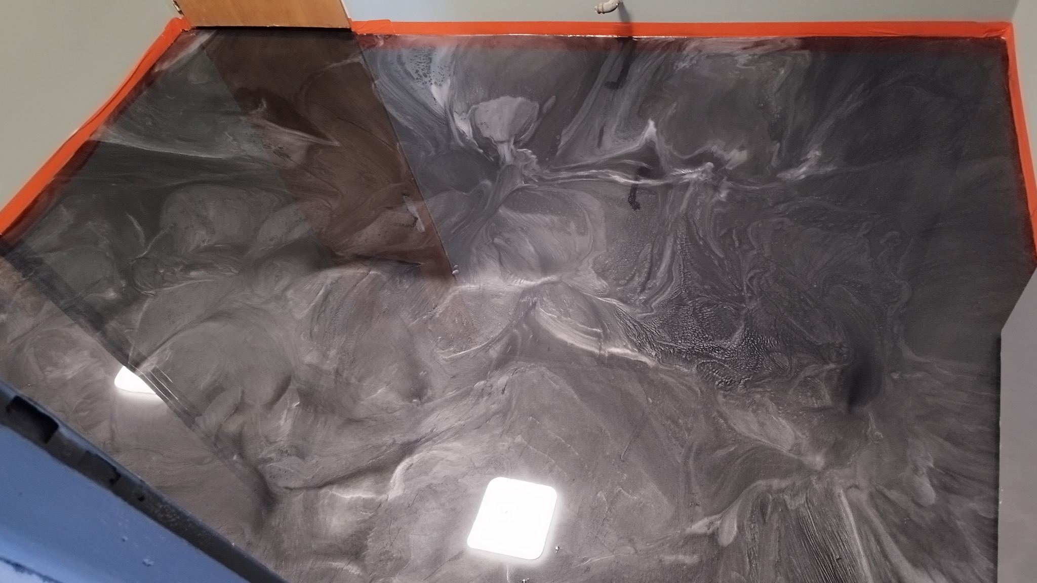 Szara, błyszcząca podłoga z efektem marmuru i pomarańczową taśmą malarską wzdłuż krawędzi ściany.