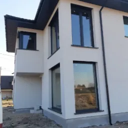 Nowoczesny, dwupiętrowy dom z jasną, gładką elewacją, czarnymi ramami okiennymi i ciemnym dachem, w trakcie prac wykończeniowych wokół budynku.