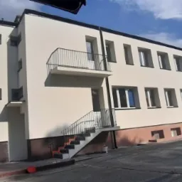 Odnowiony budynek biurowy z beżową elewacją, balkonem z czarną balustradą i schodami wejściowymi, widok z dołu na tle błękitnego nieba z chmurami.