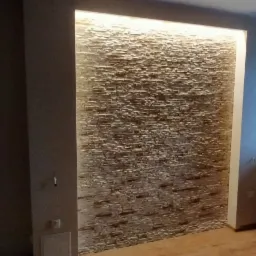 Ściana z dekoracyjnej okładziny kamiennej podświetlona ledowym światłem, widoczny fragment okna i grzejnika oraz drewniany taboret.