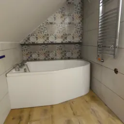 Biała wanna w łazience na poddaszu, z drewnianą podłogą i mozaiką nad wanną, grzejnik drabinkowy na ścianie