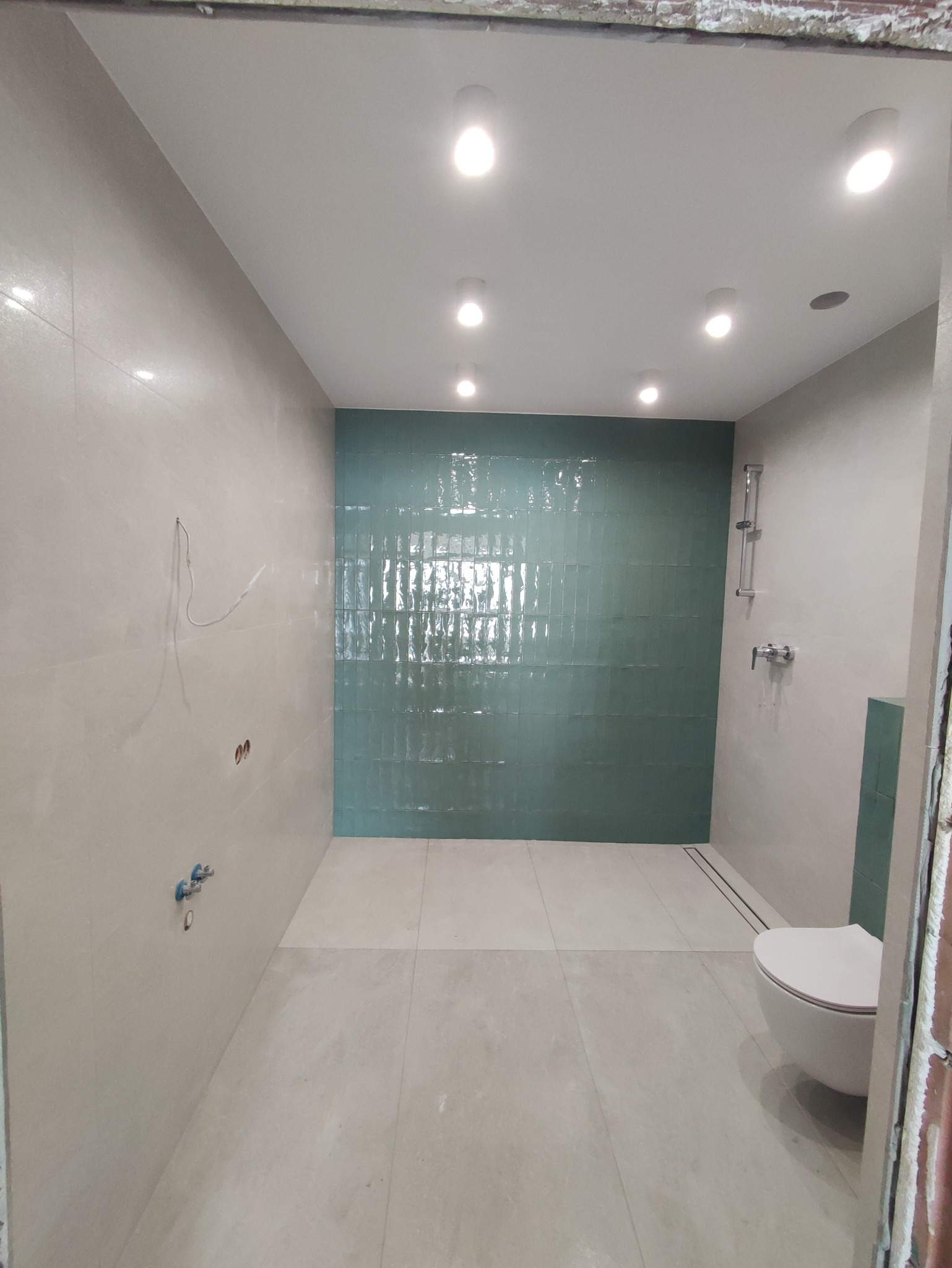 Wnętrze łazienki w trakcie remontu, widoczne turkusowe płytki na ścianie prysznicowej, biała wisząca toaleta oraz przygotowane instalacje wodno-kanalizacyjne na ścianie z szarą płytką.