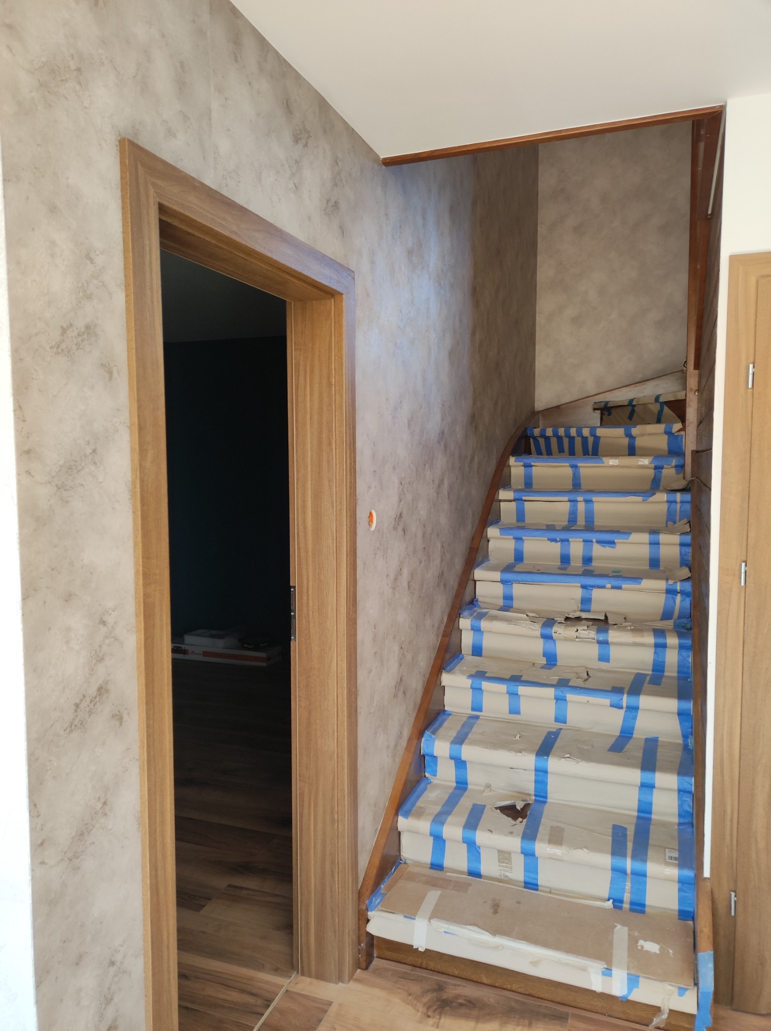Klatka schodowa zabezpieczona kartonem i niebieską taśmą malarską podczas prac wykończeniowych, widok na drewnianą futrynę drzwi i wzorzystą ścianę.
