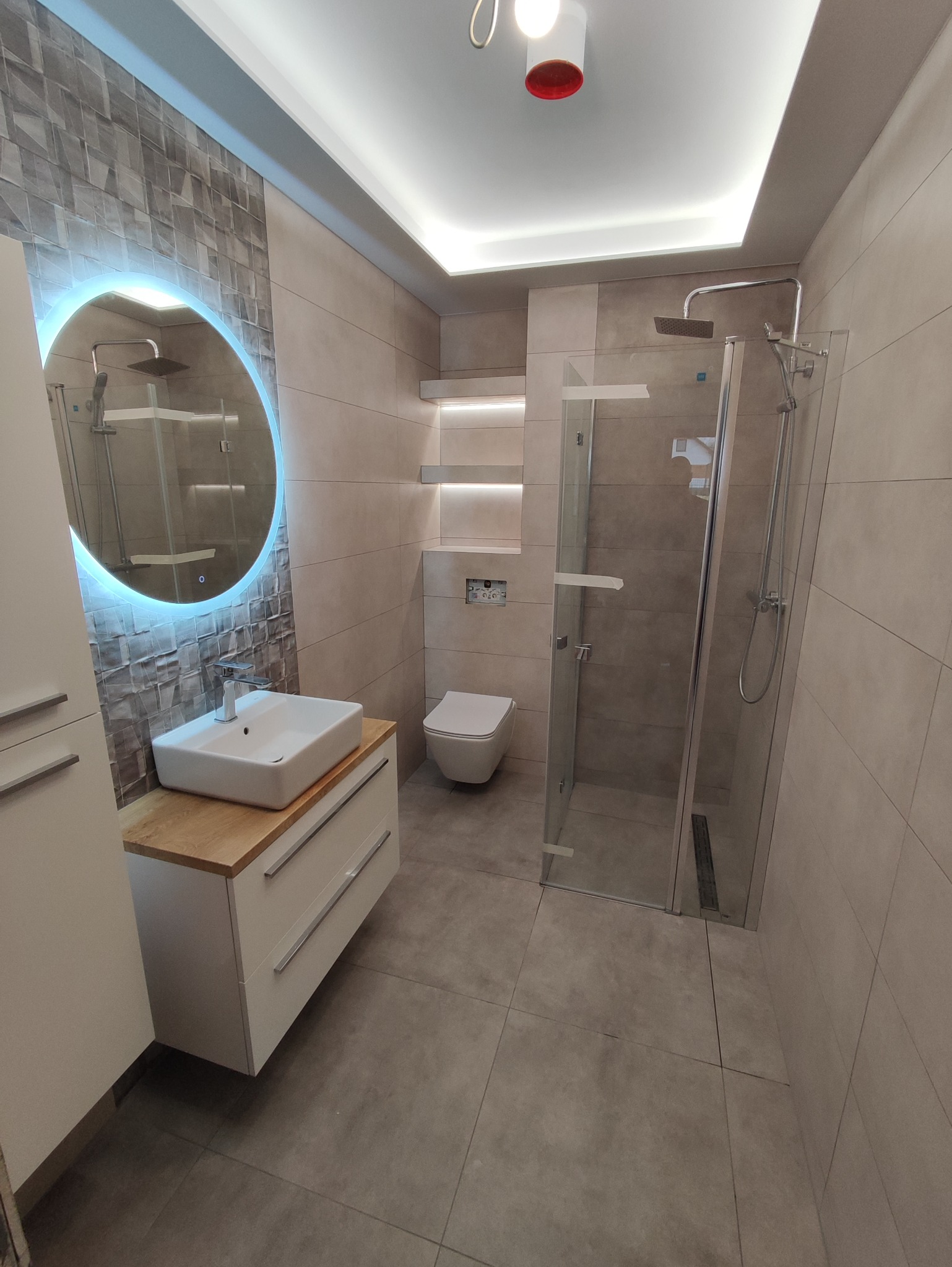 Nowoczesna łazienka z umywalką nablatową na drewnianym blacie, okrągłym lustrem z podświetleniem LED, kabiną prysznicową typu walk-in i podwieszaną toaletą.