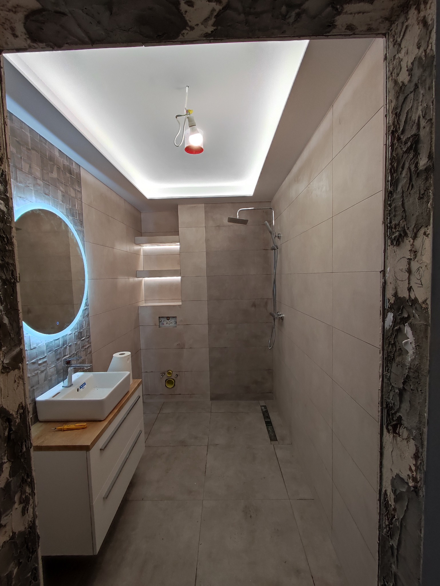 Nowoczesna łazienka z beżowymi płytkami, podświetlanym lustrem i półkami, widoczny fragment surowej ościeżnicy oraz żółty śrubokręt na blacie szafki pod umywalką.