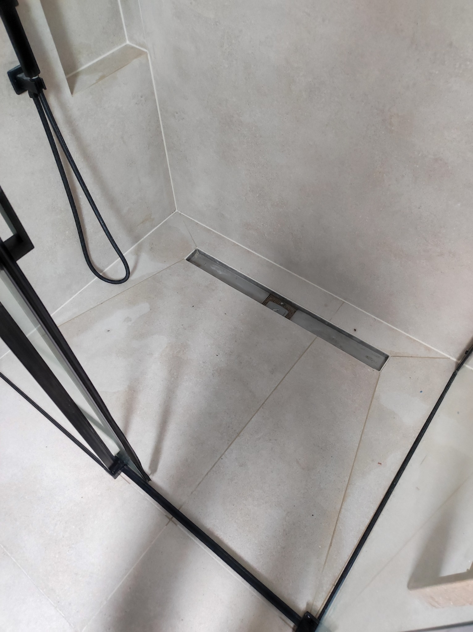 Nowoczesny odpływ liniowy w kabinie prysznicowej z szarymi płytkami, czarna armatura i szklane drzwi.