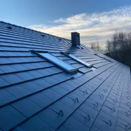 Nowy dach z ciemnej dachówki ceramicznej z zamontowanymi oknami dachowymi i kominem, widoczne zabezpieczenia przeciwśniegowe w trakcie montażu, na tle błękitnego nieba z chmurami i drzew bez liści.