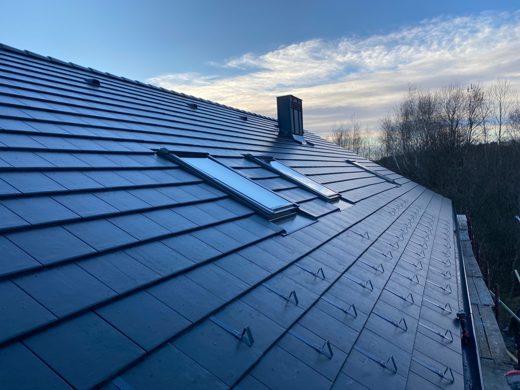Nowy dach z ciemnej dachówki ceramicznej z zamontowanymi oknami dachowymi i kominem, widoczne zabezpieczenia przeciwśniegowe w trakcie montażu, na tle błękitnego nieba z chmurami i drzew bez liści.