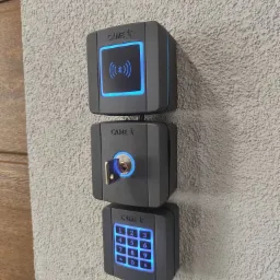 Zestaw trzech modułów kontroli dostępu CAME na szarej, chropowatej ścianie: czytnik zbliżeniowy Bluetooth, zamek na klucz i klawiatura numeryczna z niebieskim podświetleniem.