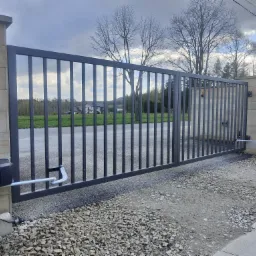 Automatyczna brama przesuwna z pionowymi szczeblami, zamontowana między kamiennymi słupkami, widoczne siłowniki, w tle domy i drzewa.