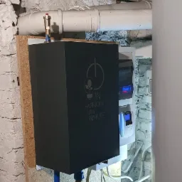 Czarna obudowa urządzenia z napisem 'In Harmony with Nature' zamontowana na ścianie z białej cegły, widoczne rury instalacyjne i panel sterowania z wyświetlaczem.