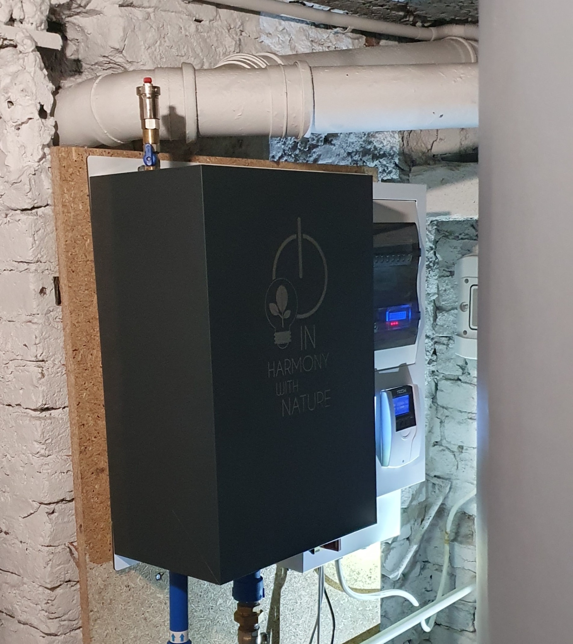 Czarna obudowa urządzenia z napisem 'In Harmony with Nature' zamontowana na ścianie z białej cegły, widoczne rury instalacyjne i panel sterowania z wyświetlaczem.