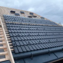 Nowy dach z czarnej dachówki ceramicznej układany na drewnianej konstrukcji więźby dachowej, widoczne fragmenty rynny i niezabezpieczone krawędzie dachu.