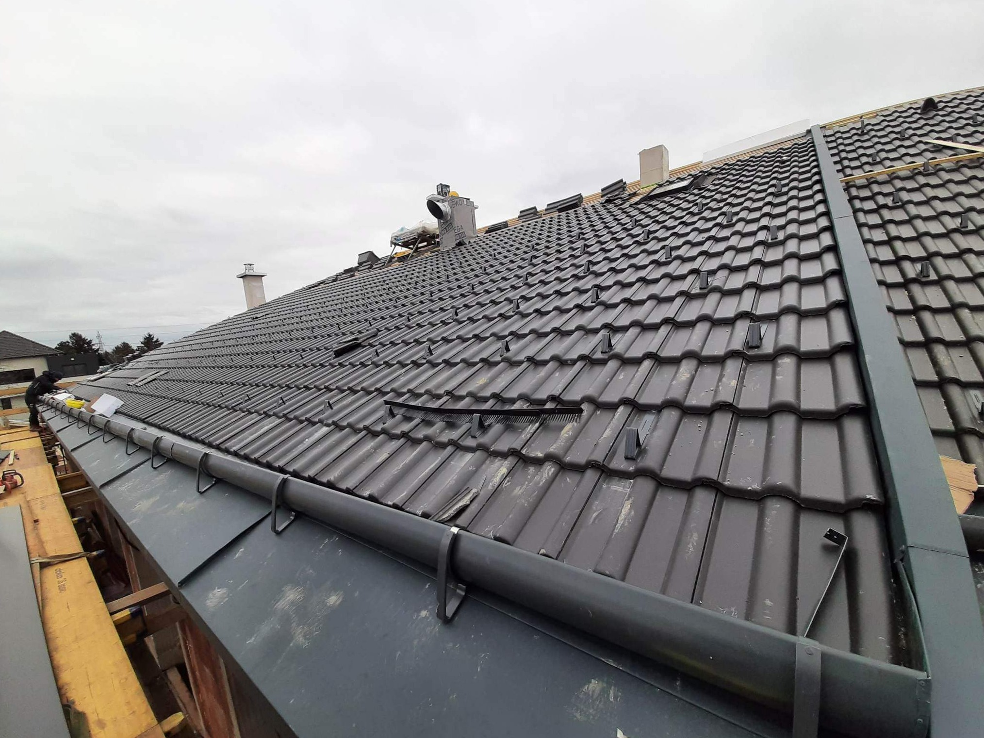 Montaż rynny na nowym, ciemnoszarym dachu z dachówki ceramicznej, widoczne elementy mocujące rynnę, fragment komina w tle, pochmurne niebo.
