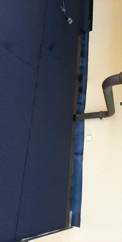 Fragment nowej, ciemnoniebieskiej papy termozgrzewalnej na dachu, widoczne łączenia i obróbki blacharskie przy ścianie budynku z rurą spustową.