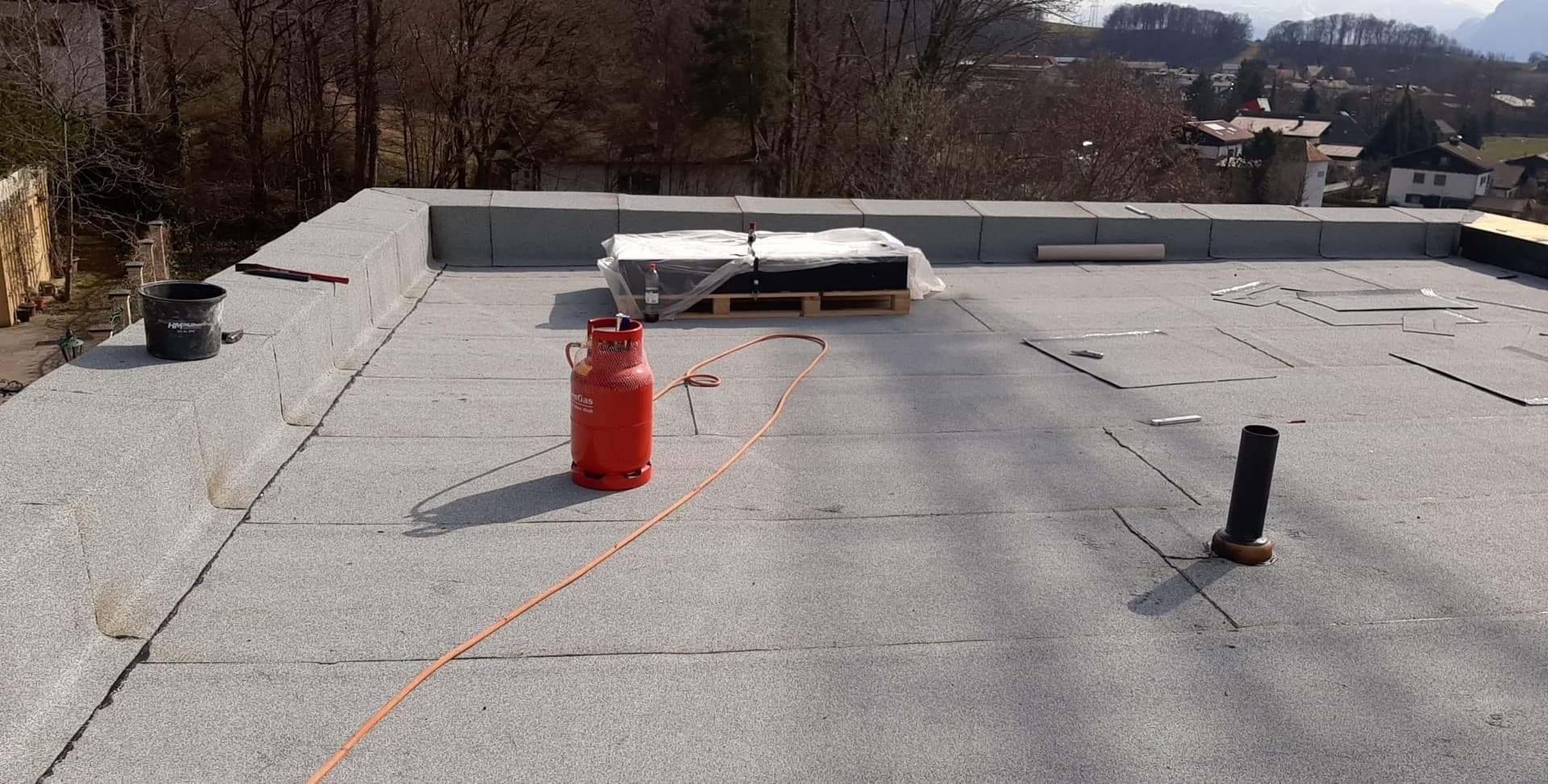 Płaski dach pokryty papą termozgrzewalną podczas prac, widoczna butla gazowa, paleta z materiałem, wiadro i komin wentylacyjny, w tle zabudowa jednorodzinna i drzewa.