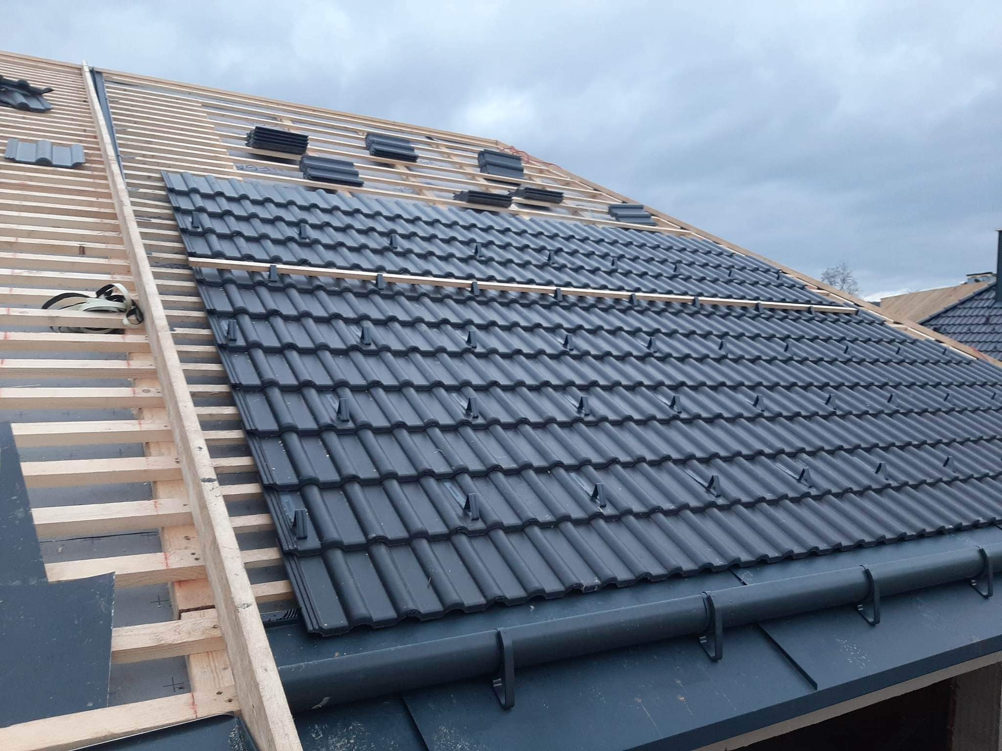 Nowy dach z czarnej dachówki ceramicznej układany na drewnianej konstrukcji więźby dachowej, widoczne fragmenty rynny i niezabezpieczone krawędzie dachu.