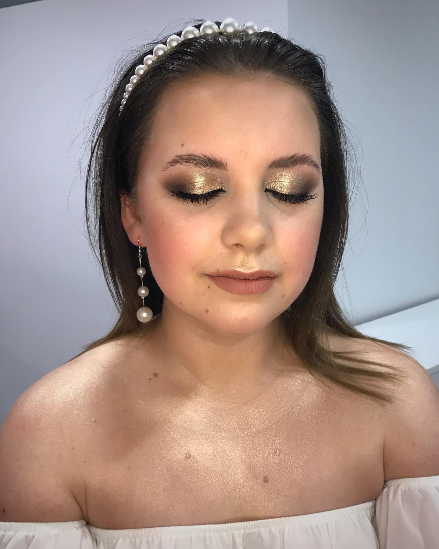 Portret kobiety z zamkniętymi oczami, prezentującej złoty makijaż oczu, matową pomadkę w odcieniu nude, perłową opaskę na włosach i długie kolczyki z pereł.