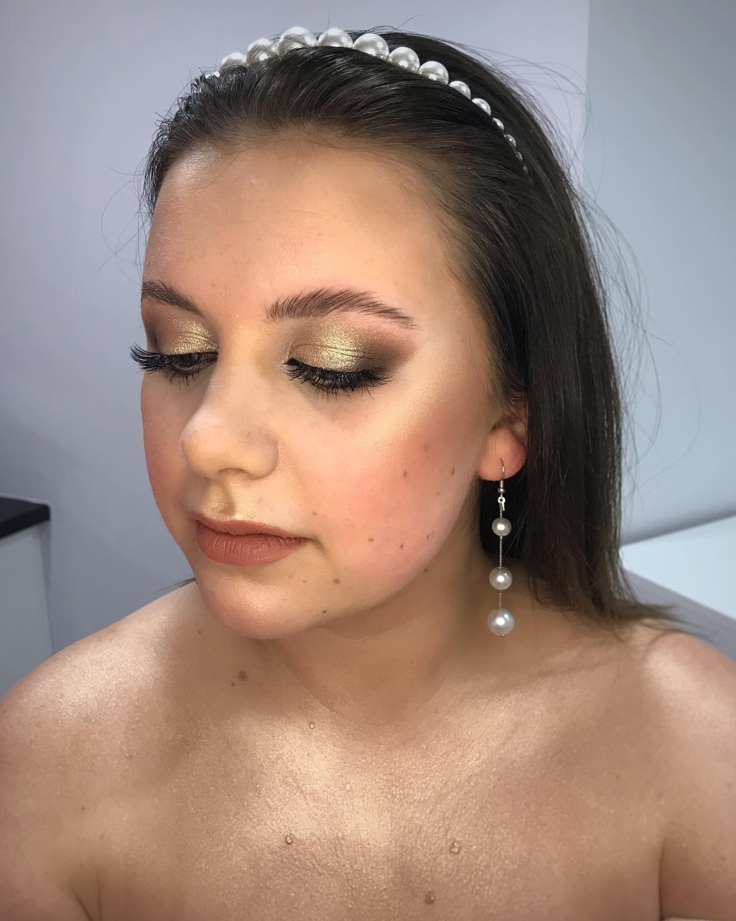 Portret kobiety z zamkniętymi oczami, prezentującej złoty makijaż oczu, podkreślone kości policzkowe, matowe usta w odcieniu nude, perłową opaskę na włosach i wiszące kolczyki z trzema perłami.