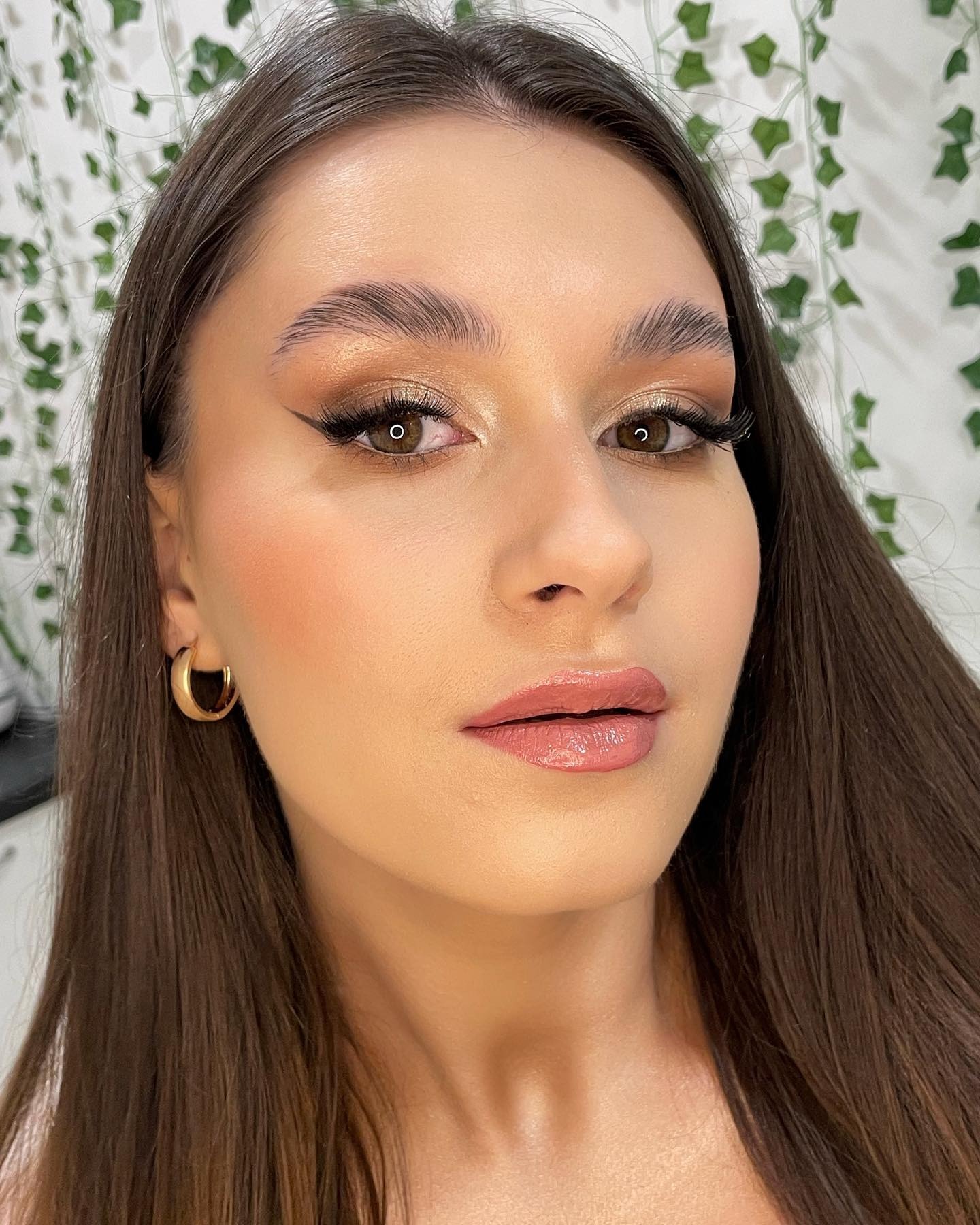 Portret kobiety z profesjonalnym makijażem: złote cienie z czarną kreską, podkreślone brwi, róż na policzkach, błyszczące usta. W tle ściana z motywem roślinnym.