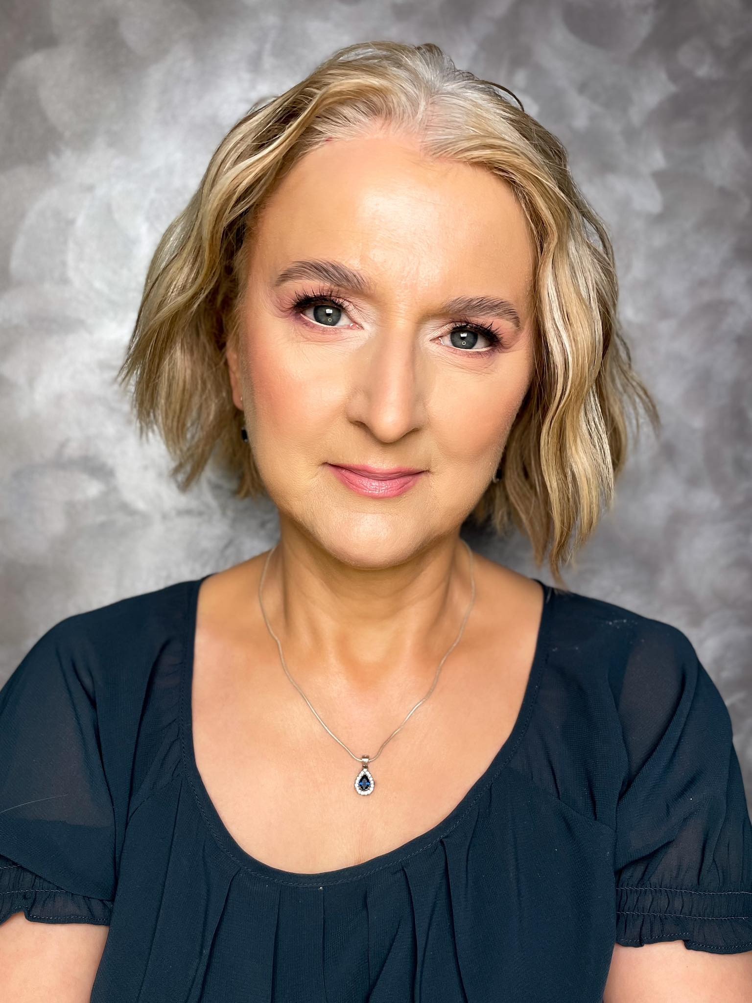 Portret kobiety z delikatnym makijażem, blond włosami z odrostami i srebrnym naszyjnikiem na tle szarego, teksturowanego tła.