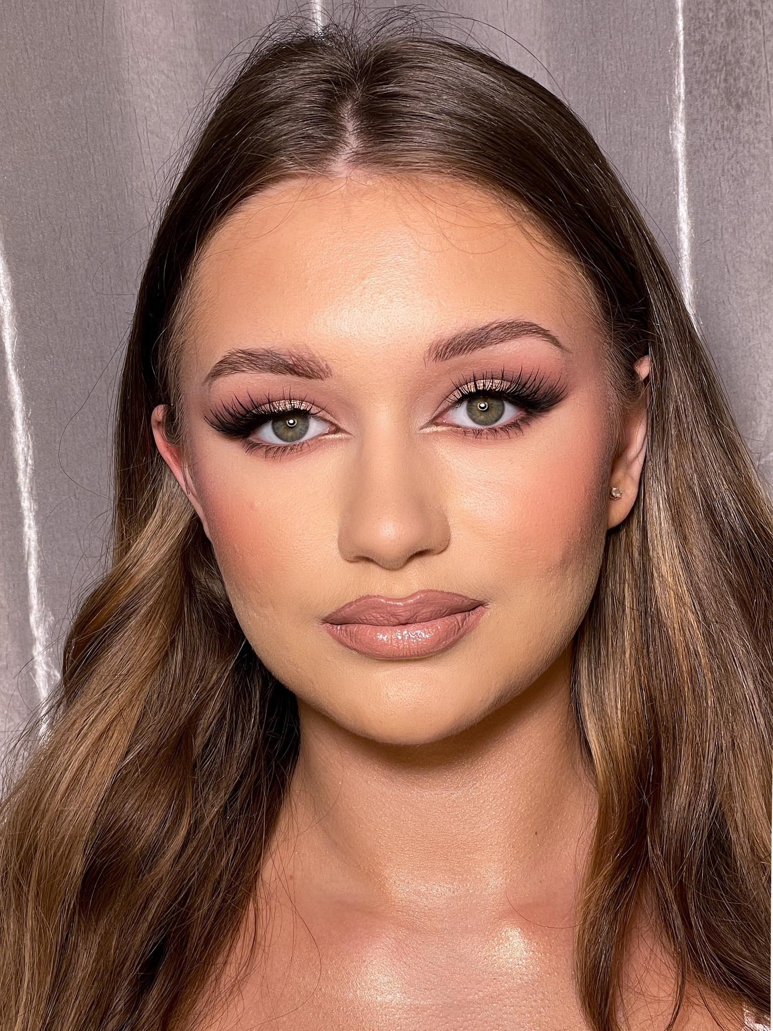 Portret kobiety z profesjonalnym makijażem: podkreślone brwi, sztuczne rzęsy, błyszczące usta w odcieniu nude i delikatny róż na policzkach, z kolczykiem w uchu na tle srebrnej zasłony.