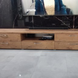 Zakład MEBLARSKI Meblostyl - Drewniana szafka RTV z szufladami i otwartą półką, stojąca na jasnej podłodze, pod telewizorem na tle ściany z czarnego marmuru.