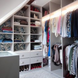 Zakład MEBLARSKI Meblostyl - Biała szafa na wymiar zabudowana na poddaszu ze skośnym sufitem, z oświetleniem LED i dekoracyjnym wzorem na tylnej ściance półek.