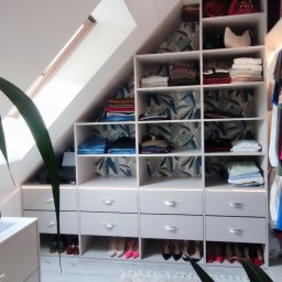 Zakład MEBLARSKI Meblostyl - Biała szafa wnękowa na poddaszu ze skośnym sufitem, z półkami wypełnionymi ubraniami i szufladami z dekoracyjnymi uchwytami. U dołu rząd butów.