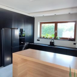 Zakład MEBLARSKI Meblostyl - Nowoczesna kuchnia: czarne szafki, drewniany blat, okno z roślinami. Minimalistyczny design z akcentem naturalnego drewna i świeżej zieleni. Funkcjonalność i estetyka.