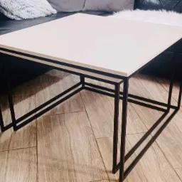 Nowoczesny stolik kawowy z białym blatem i czarnymi, metalowymi nogami o geometrycznym designie, ustawiony na podłodze imitującej drewno.