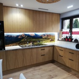 Zakład MEBLARSKI Meblostyl - Jasna kuchnia z drewnianymi frontami i blatem w odcieniach szarości. Nad blatem panel z motywem górskim. Na suficie dekoracyjny, okrągły element oświetleniowy.