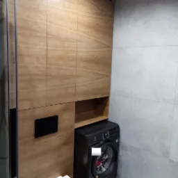 Nowoczesna łazienka z drewnianą zabudową meblową, podwieszaną toaletą i czarną pralką. Minimalistyczny design z szarymi płytkami.
