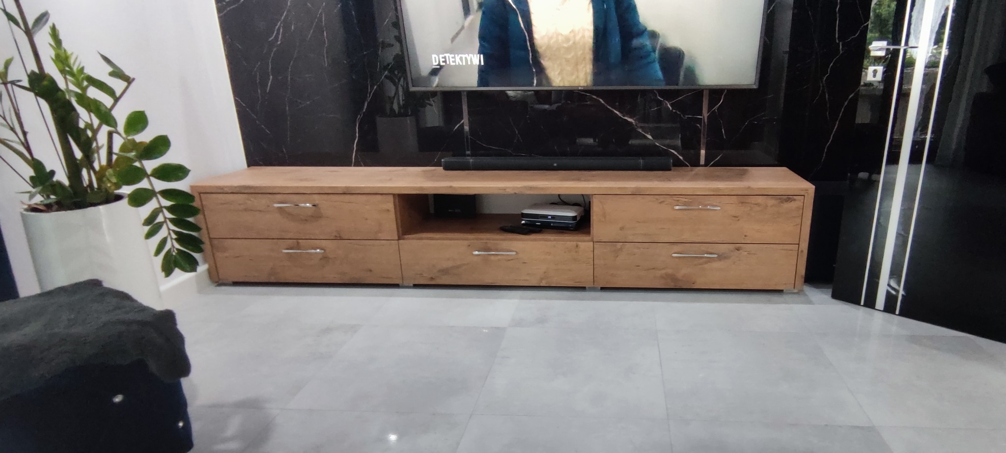 Drewniana szafka RTV z szufladami i otwartą półką, stojąca na jasnej podłodze, pod telewizorem na tle ściany z czarnego marmuru.