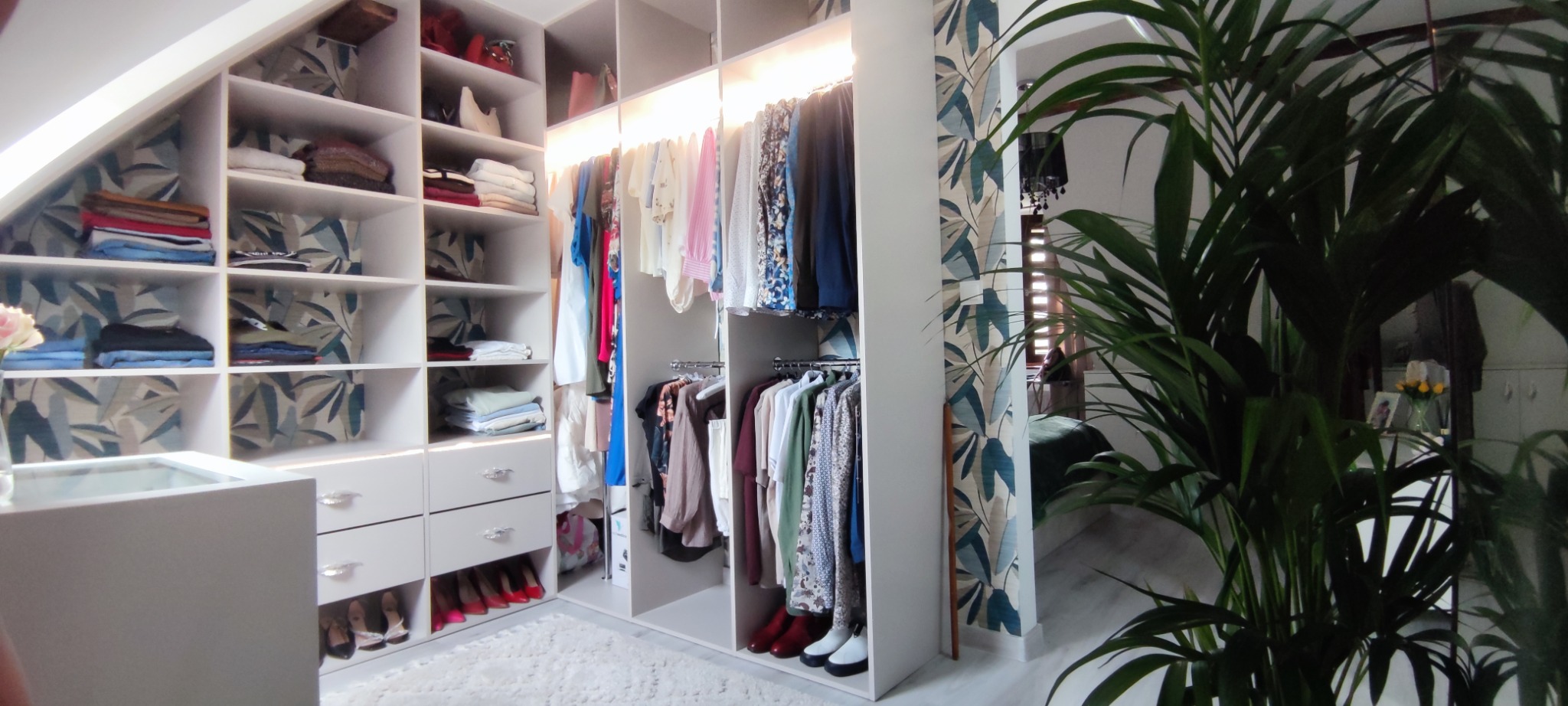 Biała garderoba na poddaszu z półkami, szufladami i wieszakami, oświetlona listwami LED, z wzorzystą tapetą w tle i rośliną doniczkową na pierwszym planie.