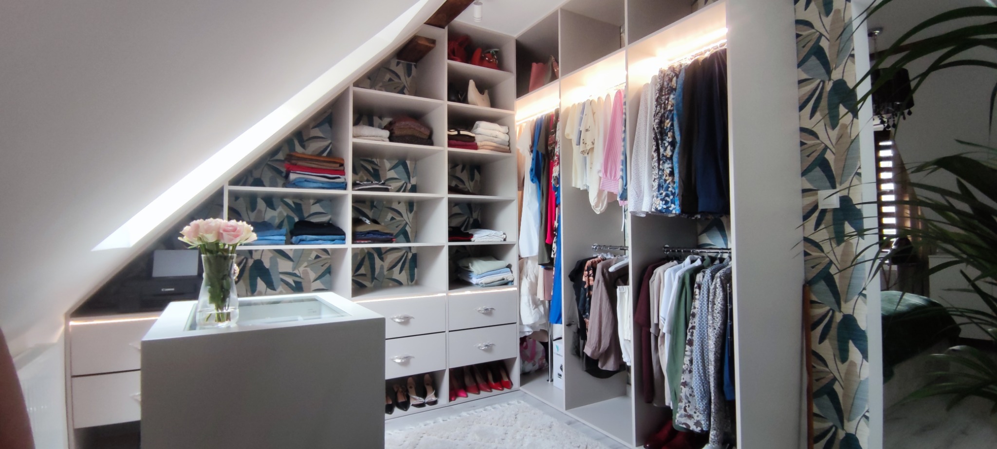 Biała szafa na wymiar zabudowana na poddaszu ze skośnym sufitem, z oświetleniem LED i dekoracyjnym wzorem na tylnej ściance półek.