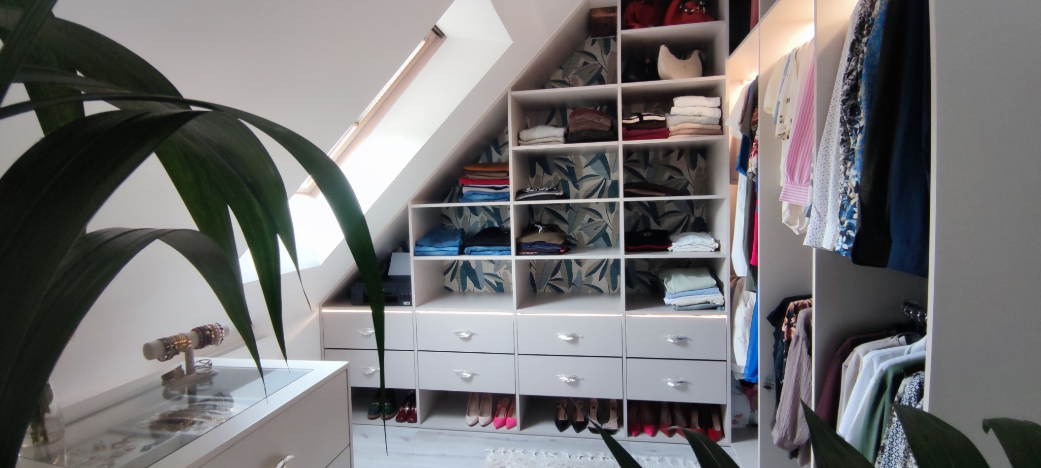 Biała szafa wnękowa na poddaszu ze skośnym sufitem, z półkami wypełnionymi ubraniami i szufladami z dekoracyjnymi uchwytami. U dołu rząd butów.