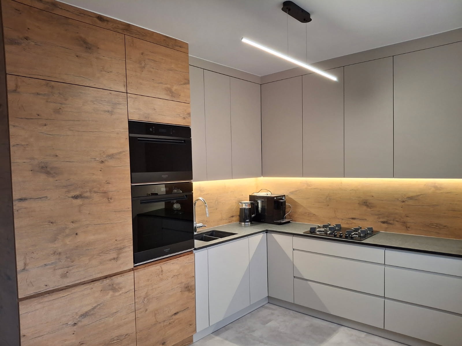 Nowoczesna kuchnia w odcieniach szarości i drewna z oświetleniem LED, zabudowana piekarnikiem i płytą gazową, z ekspres do kawy na blacie.