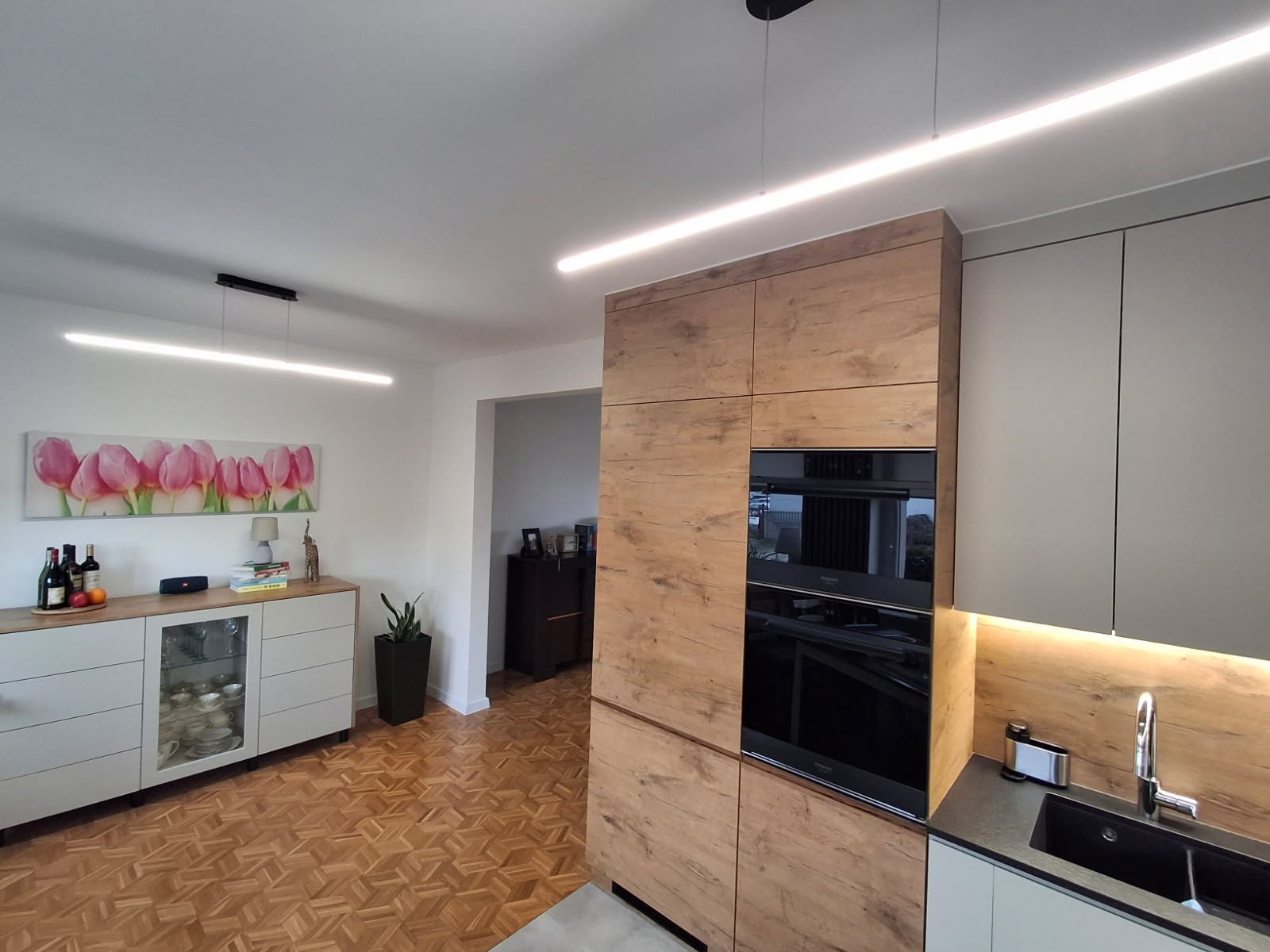 Nowoczesna kuchnia: zabudowa z drewnopodobnym frontem, wbudowany piekarnik, szare szafki, oświetlenie LED i parkiet w jodełkę tworzą spójną całość.